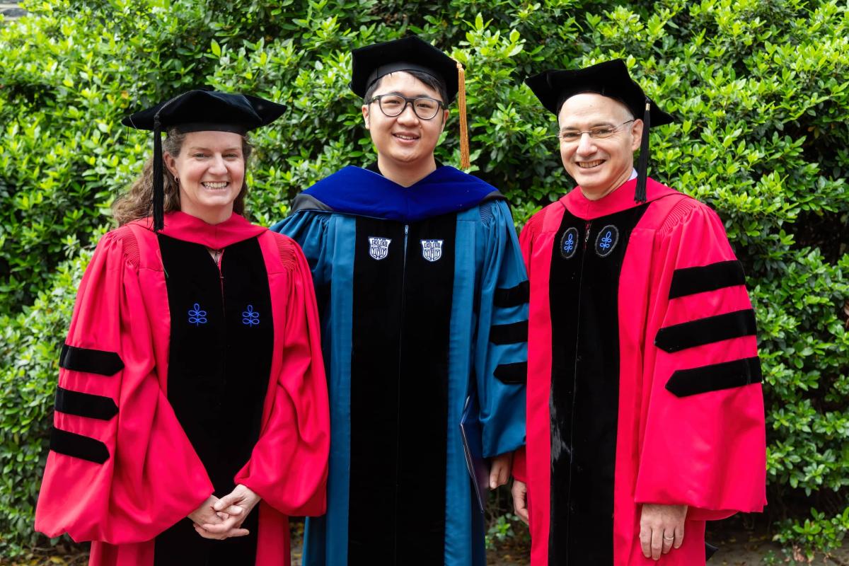 Man in blue cap and gown standing with two professors in red.