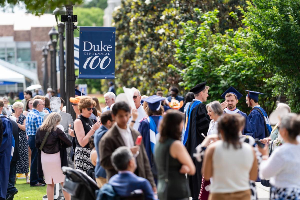 Duke 100 sign, with people in foreground, milling around