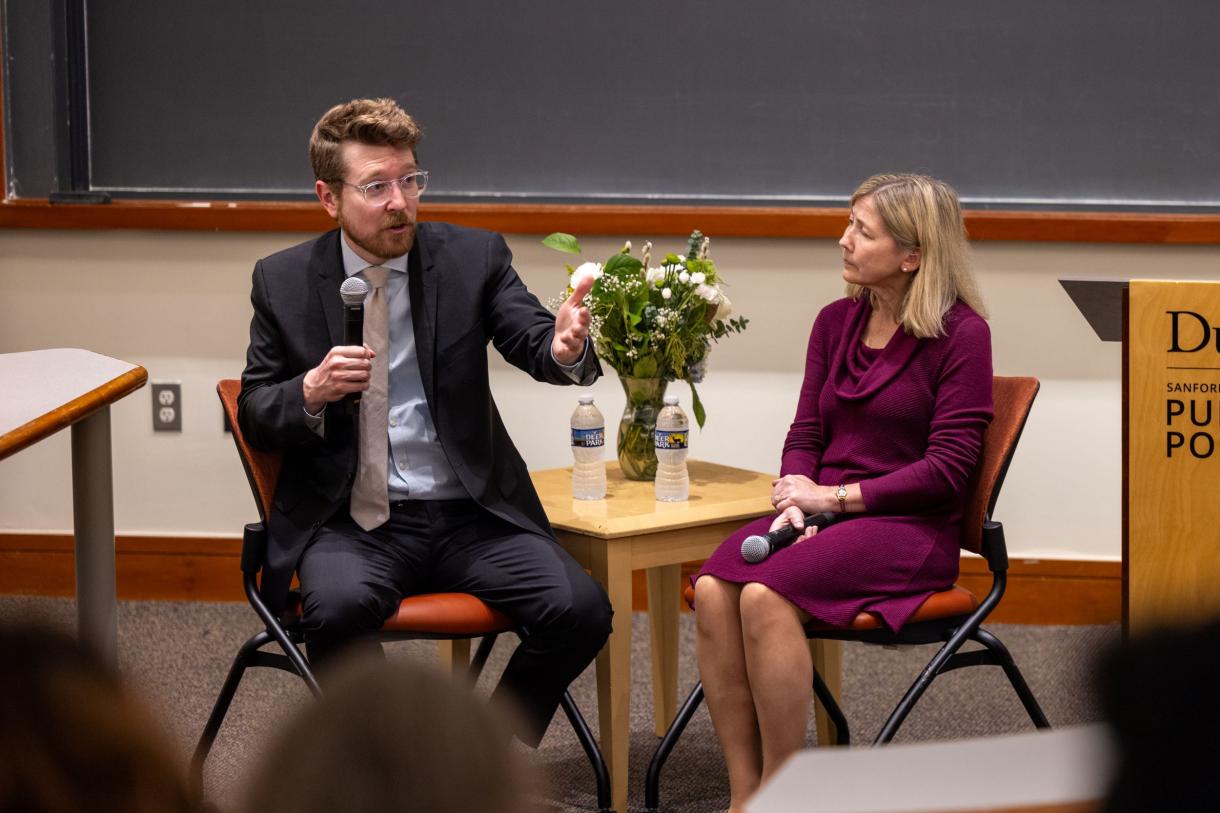 Man in suit gesturing towards person in crowd while he speaks and woman moderator on right looks on. 