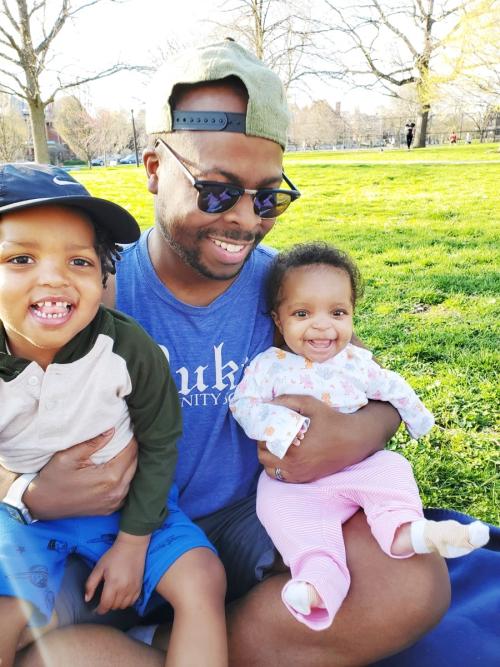 Man wearing Duke shirt while holding two small children. 