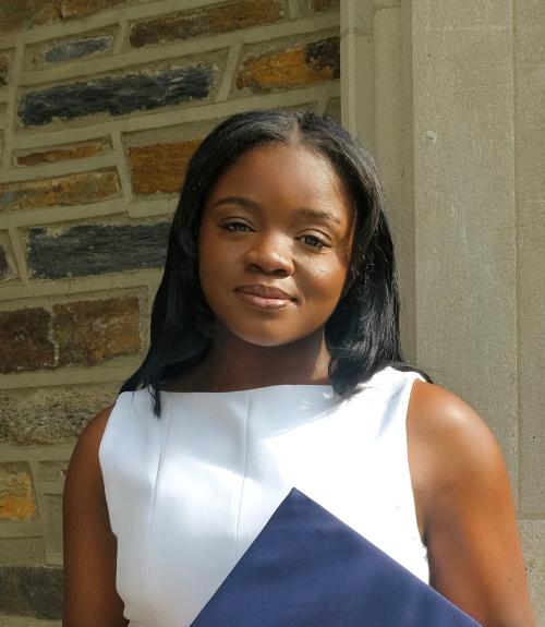 Bunmi Omisore smiling while standing in front of a brick wall and holding a blue graduation cap. 